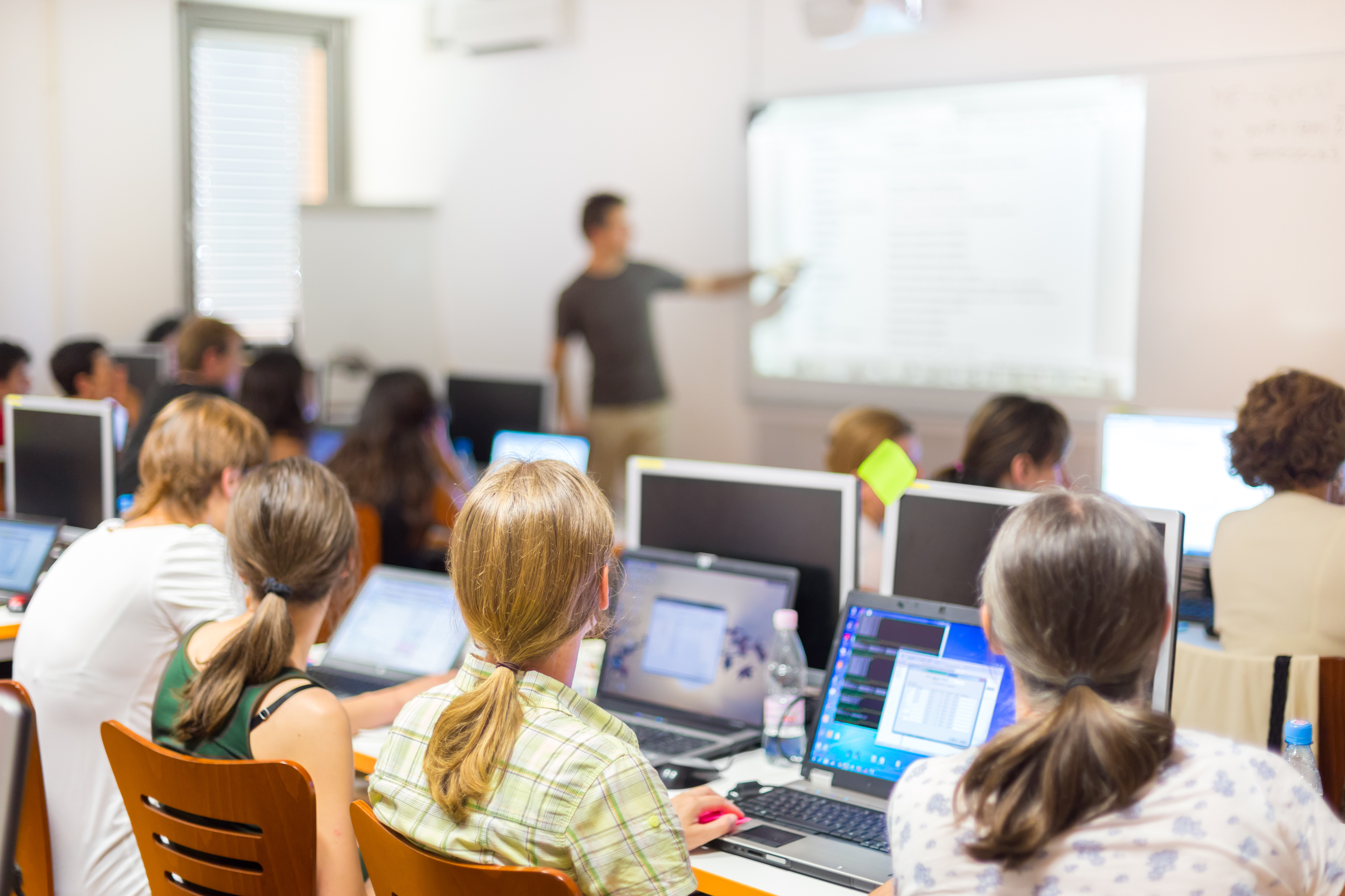 An image of a man pointing to a whiteboard giving a presentation to students sat at their desks